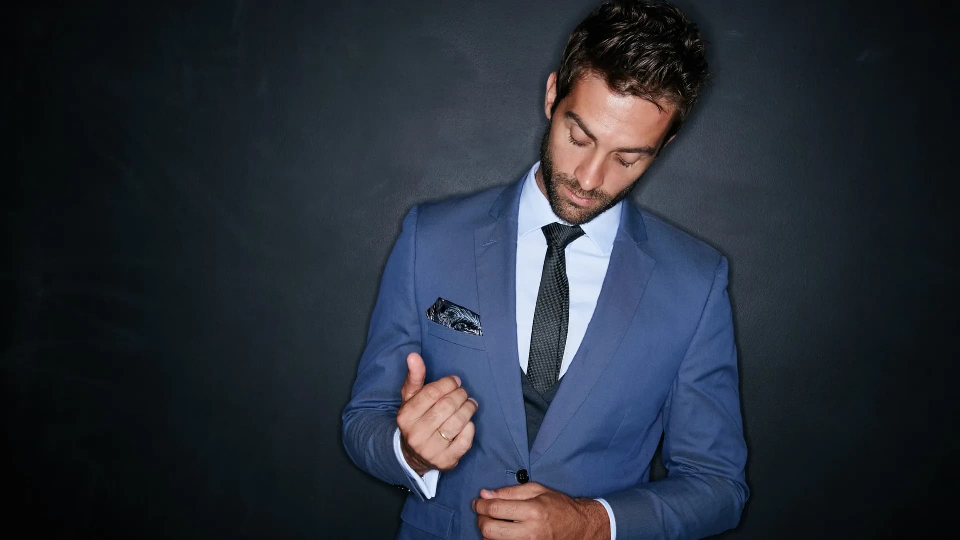 Man in a blue suit adjusting his cuff against a dark wall
